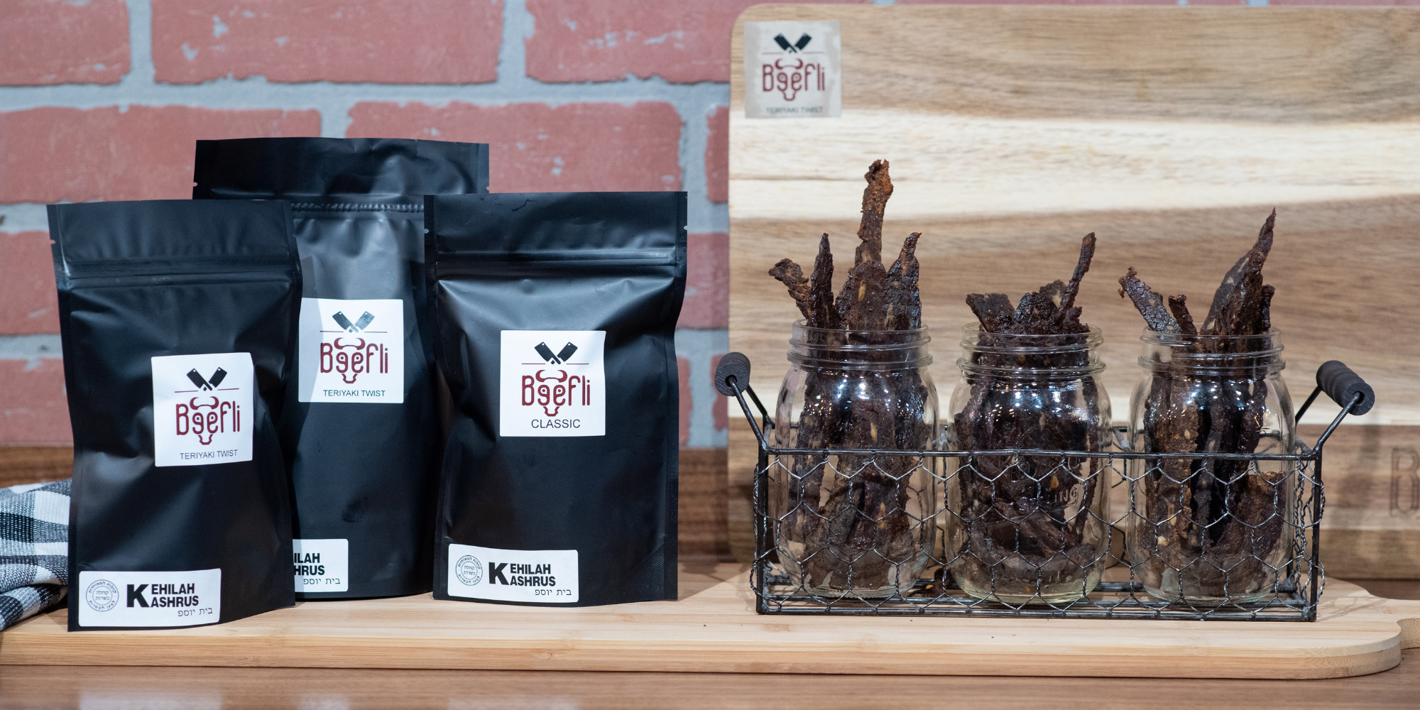 Beefli kosher gourmet beef jerky display featuring Teriyaki Twist and Classic flavors in black pouches and glass jars, set on a wooden board with a brick wall background.