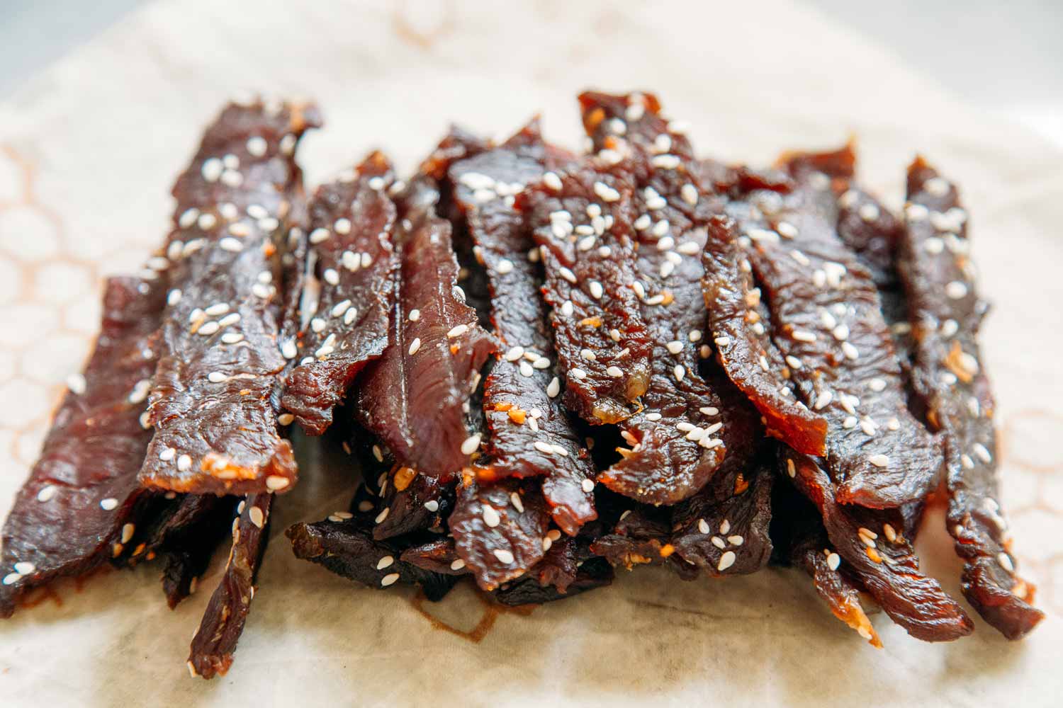 Close-up of teriyaki beef jerky strips topped with sesame seeds on parchment paper, showcasing a glossy, savory texture.